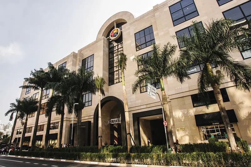 Fachada de edifício comercial em tom bege com palmeiras e grandes janelas.