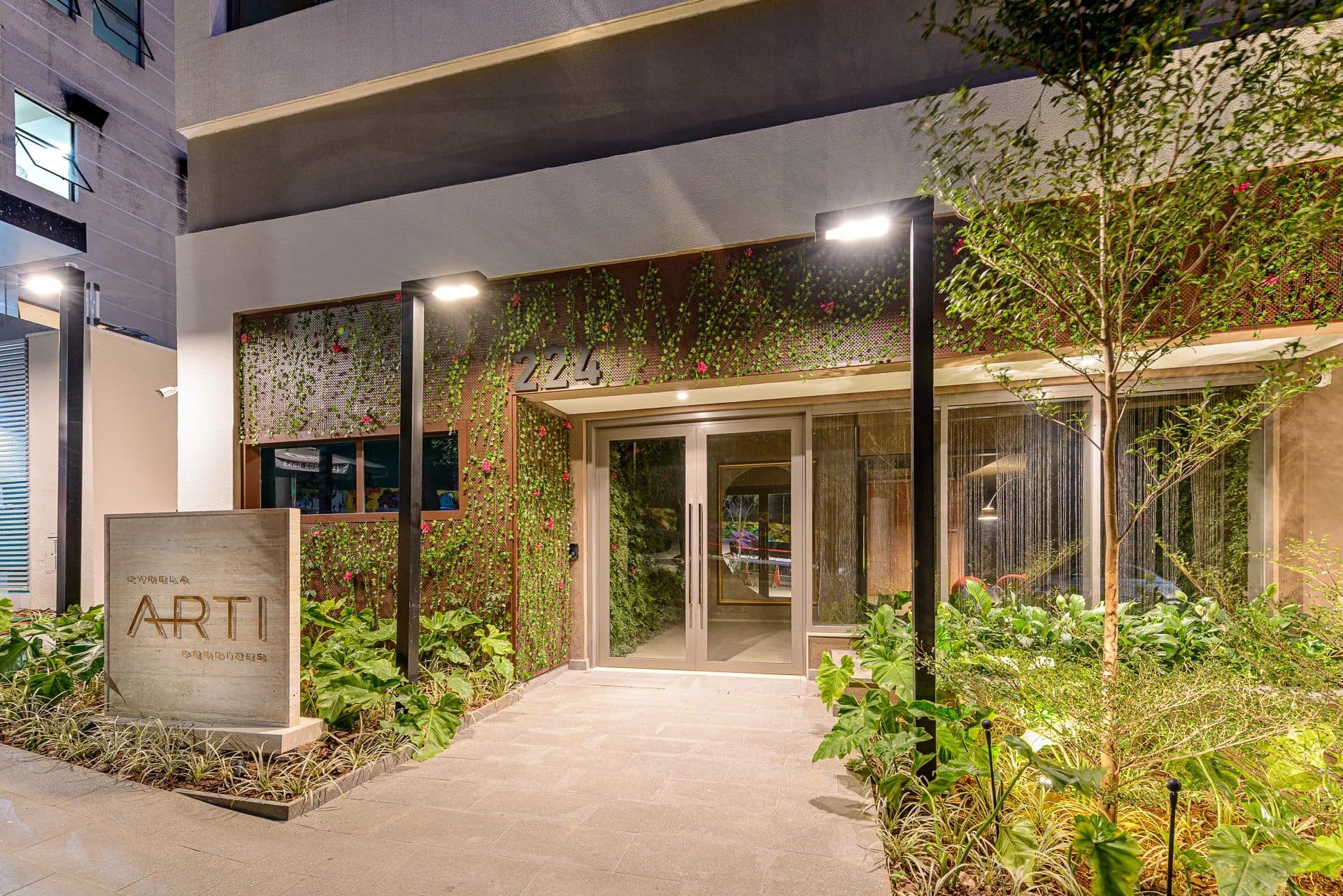 Entrada do edifício com portaria de vidro, vegetação na fachada e iluminação embutida.