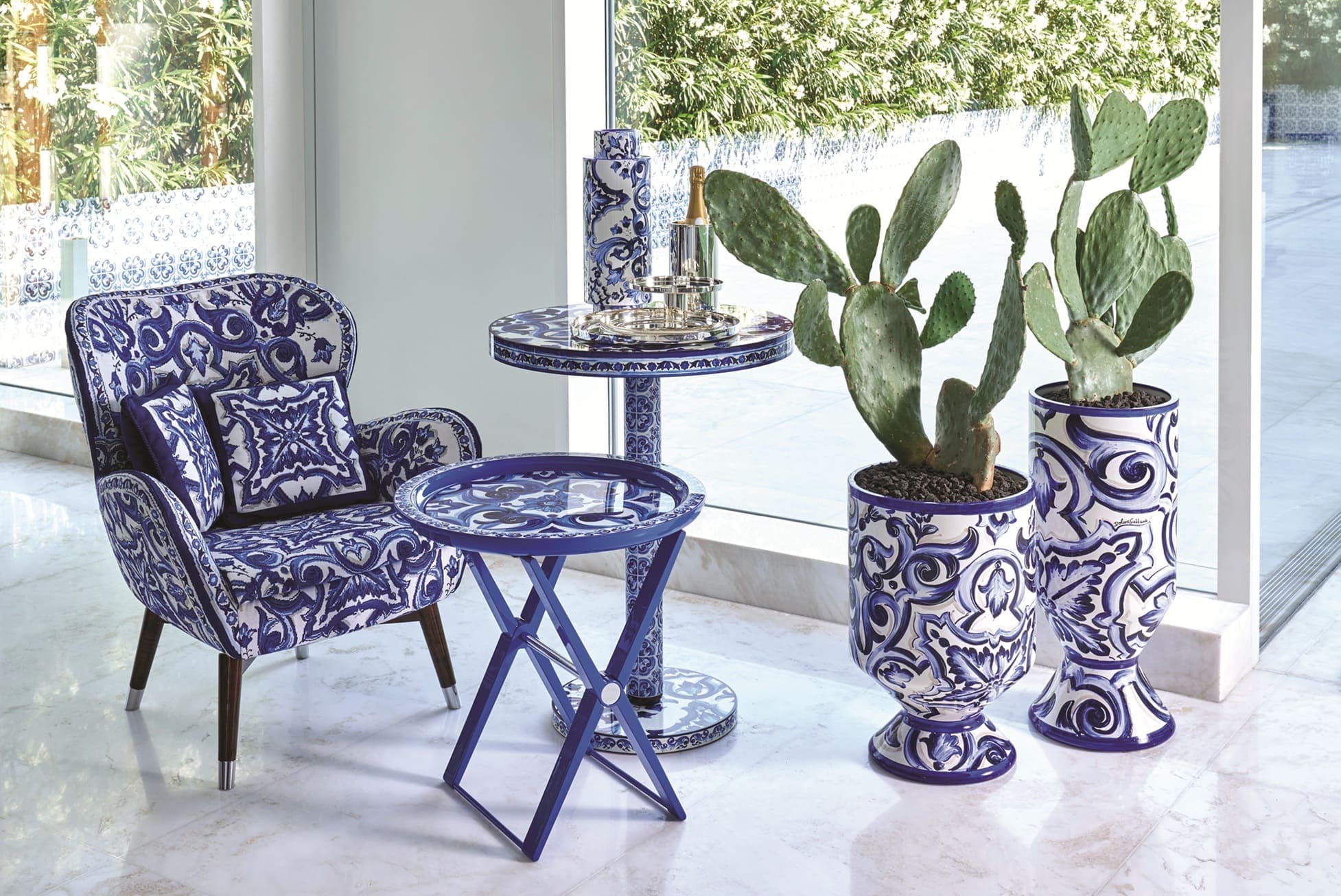 Ambiente interno com poltrona estampada em azul e branco, mesas de apoio e vasos com cactos.