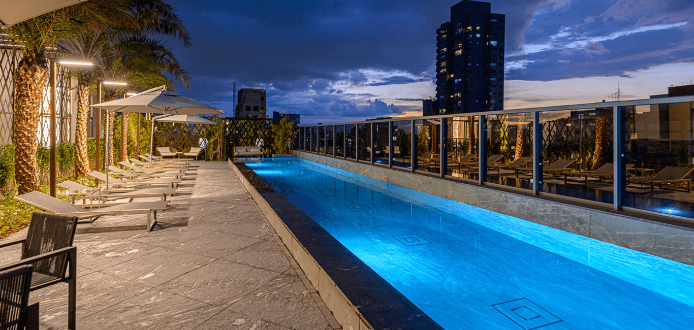 Piscina iluminada no rooftop com espreguiçadeiras e vista para a cidade ao entardecer.